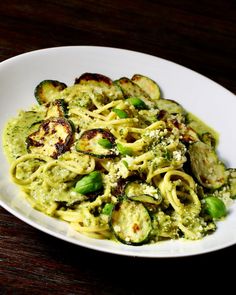 a white bowl filled with pasta and vegetables on top of a wooden table next to a fork