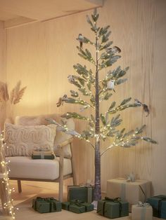 a small christmas tree with presents under it in front of a couch and wall decorated with lights