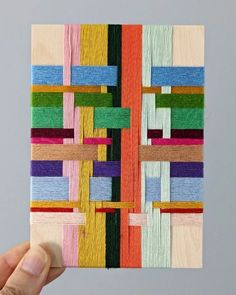 a hand holding up a piece of art made out of strips of wood and yarn