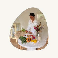 a woman in white shirt cutting vegetables on table with vases and flowers behind her