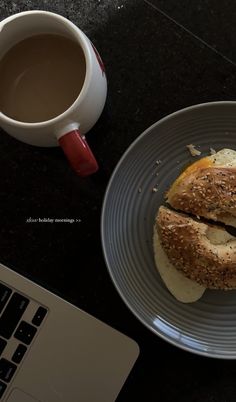 a cup of coffee next to a plate with two sandwiches on it and a laptop