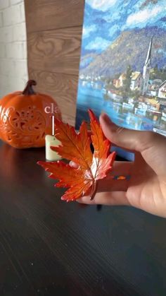 a hand holding an orange maple leaf next to a small pumpkin on a table with a painting in the background