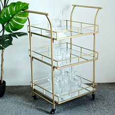 a gold bar cart filled with glasses next to a potted green plant on the floor