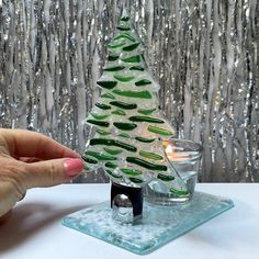 a hand holding a glass christmas tree on top of a table next to a candle