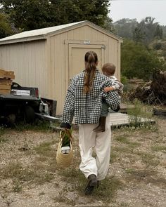 a woman carrying a child in her arms while walking towards a small building with a shed