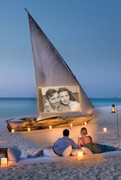 two people sitting on the beach with candles in front of them and a sailboat