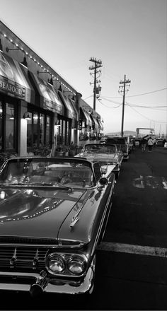 black and white photograph of classic cars parked in front of a store on the street