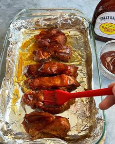 a person holding a brush over some chicken wings in a pan with sauce on the side