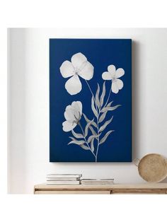 a blue painting with white flowers on the wall above a wooden table and bookcase