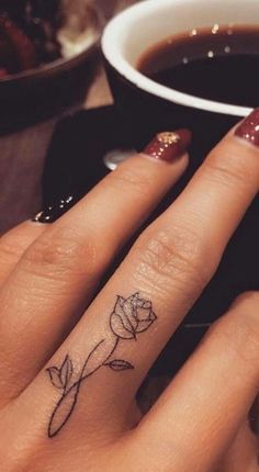 a woman's hand with a small rose tattoo on her left ring finger next to a cup of coffee