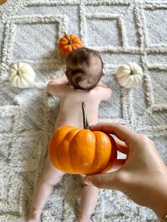 a baby is laying on the floor with a pumpkin in it's hand, while someone holds an orange object up to their face