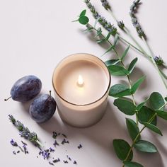 a candle and some flowers on a white surface with leaves, lavenders and two plums