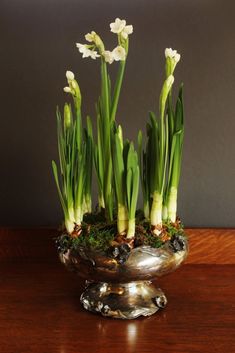 some white flowers are in a silver bowl on a wooden table with moss growing out of it