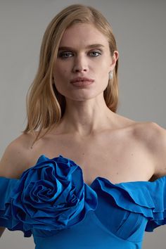 a woman wearing a blue dress with a rose on the front and shoulder, standing in front of a gray background