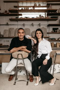 a man and woman sitting next to each other in front of shelves filled with pottery