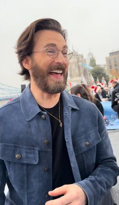 a man wearing glasses and a denim jacket smiles at the camera while standing in front of a crowd