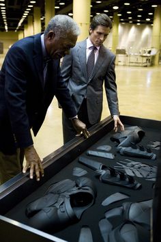 two men in suits looking at some shoes on a display case with other items behind them