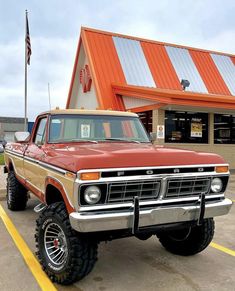 an old red and tan truck parked in front of a building next to a flag pole