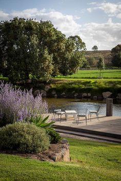 there is a table and chairs on the deck by the water with purple flowers in the foreground