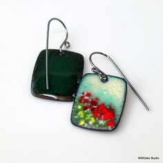 two square shaped earrings with green and red flowers in them on a white table top