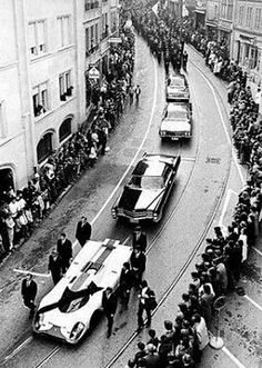 an old black and white photo of cars driving down the street with people watching them