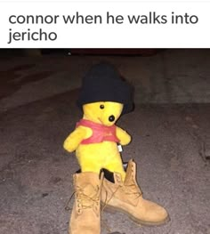 a yellow teddy bear sitting on the ground wearing boots and a hat with a red ribbon around it's neck