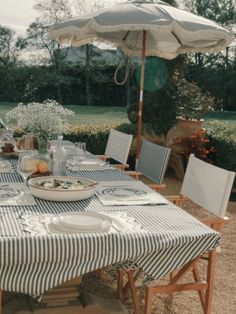 an outdoor dining table set with plates and utensils under an umbrella in the garden