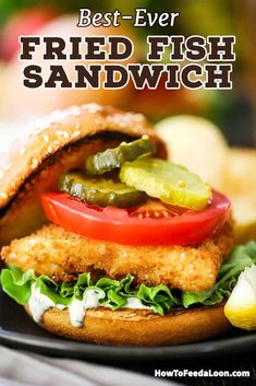 fried fish sandwich with pickles and tomatoes on a black plate text overlay reads best - ever fried fish sandwich