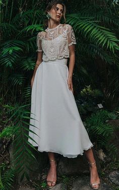 a woman wearing a white dress standing in front of some green plants and trees with her hands on her hips