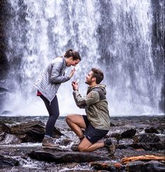 a man kneeling down next to a woman in front of a waterfall with her hands together