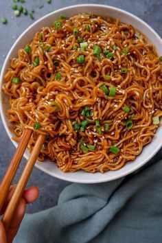 someone holding chopsticks over a bowl of noodles with sauce and scallions