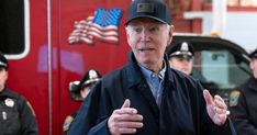 an older man standing in front of a fire truck talking to the camera with other uniformed men behind him