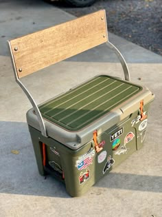 a green cooler sitting on top of a sidewalk next to a wooden bench with stickers all over it