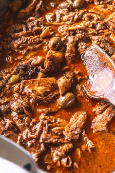 a large pot filled with meat and olives next to a wooden spoon on top of it