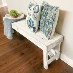 a wooden bench with two pillows and a plant on it next to a white wall