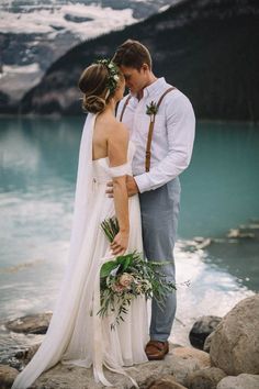 a bride and groom standing next to each other on rocks near the water with mountains in the background