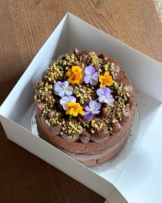 a chocolate cake with flowers on top in a white cardboard box sitting on a wooden table