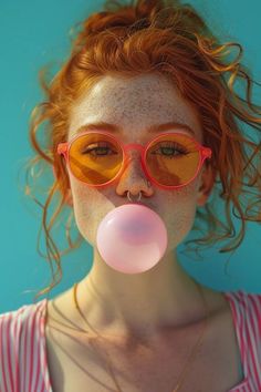 a woman with red hair and glasses blowing a bubble on her nose in front of a blue background