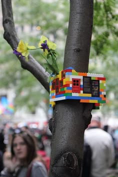 a small house made out of legos on a tree in a park with people walking around