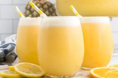 three glasses filled with orange juice next to sliced lemons and pineapple on a table