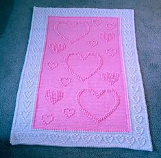 a pink and white crocheted afghan with hearts on the front, sitting on a blue carpet