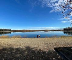 two people standing on the shore of a lake with trees in the background and blue sky