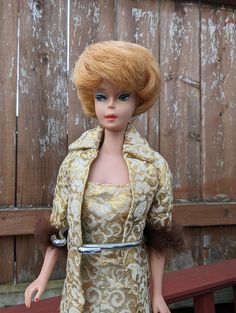 a doll with blonde hair wearing a gold dress and fur collar, standing in front of a wooden fence