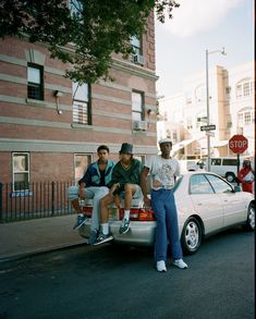 three men sitting on the back of a white car in front of a brick building