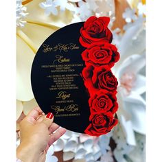 a hand holding a black and red wedding card with roses on it, in front of white flowers