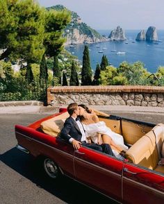 a man and woman sitting in the back of a red convertible car with an ocean view