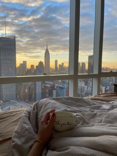 a woman laying in bed with a cup on her lap and looking out the window