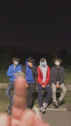 four people sitting on a bench wearing face masks at night, with one pointing to the camera