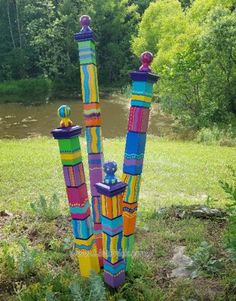 three colorful sculptures are in the grass near some water and trees, one is painted with bright colors