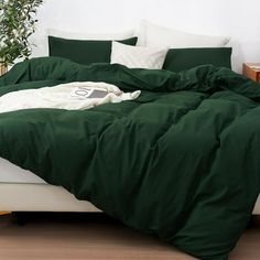 an unmade bed with green sheets and pillows in a white walled room next to a potted plant